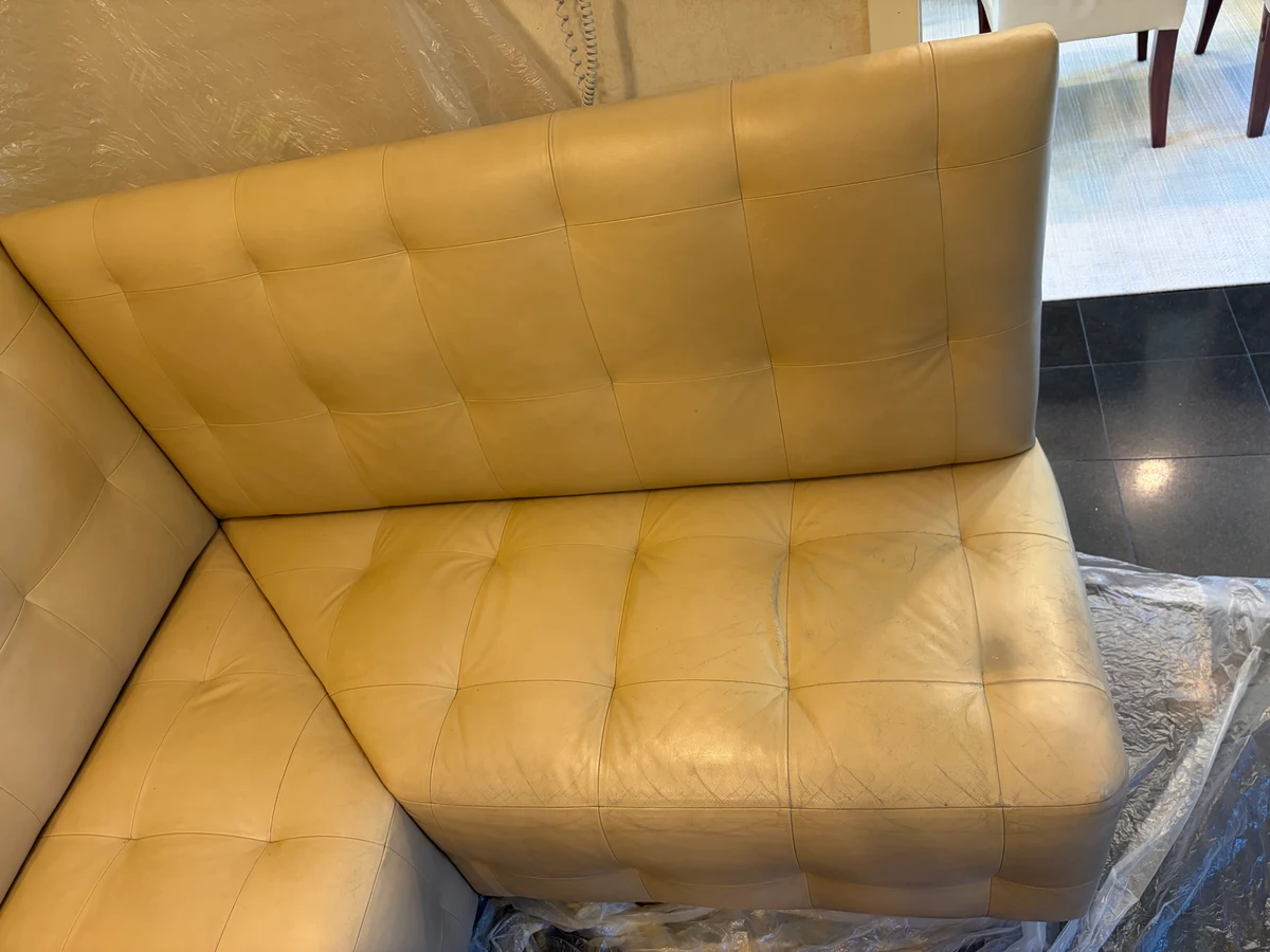 Built-in cream leather kitchen banquette corner before restoration — yellowed surface, visible cracking across seat panels, full L-shaped unit showing extent of wear