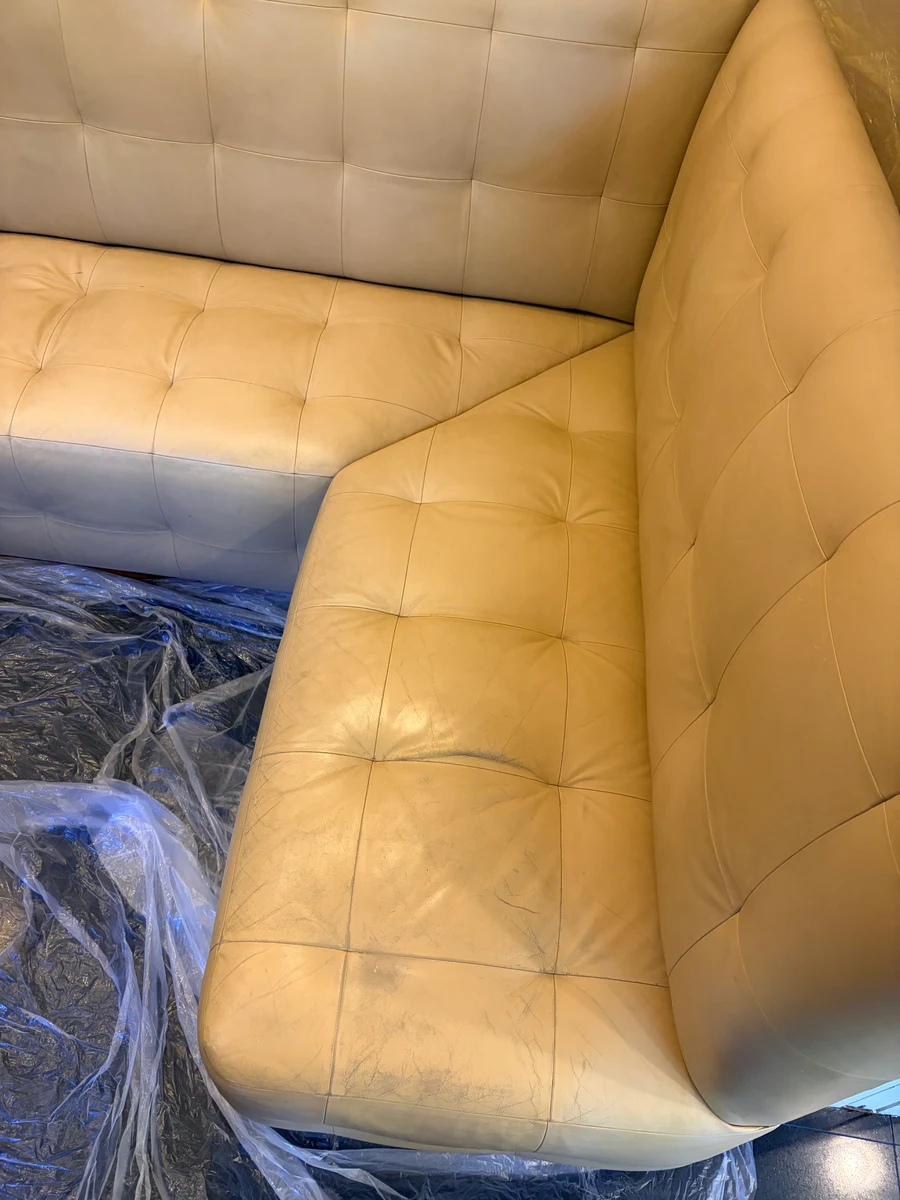 Built-in leather kitchen banquette during restoration preparation — full L-shaped corner showing contrast between cleaned and untreated sections, protective plastic sheeting on floor