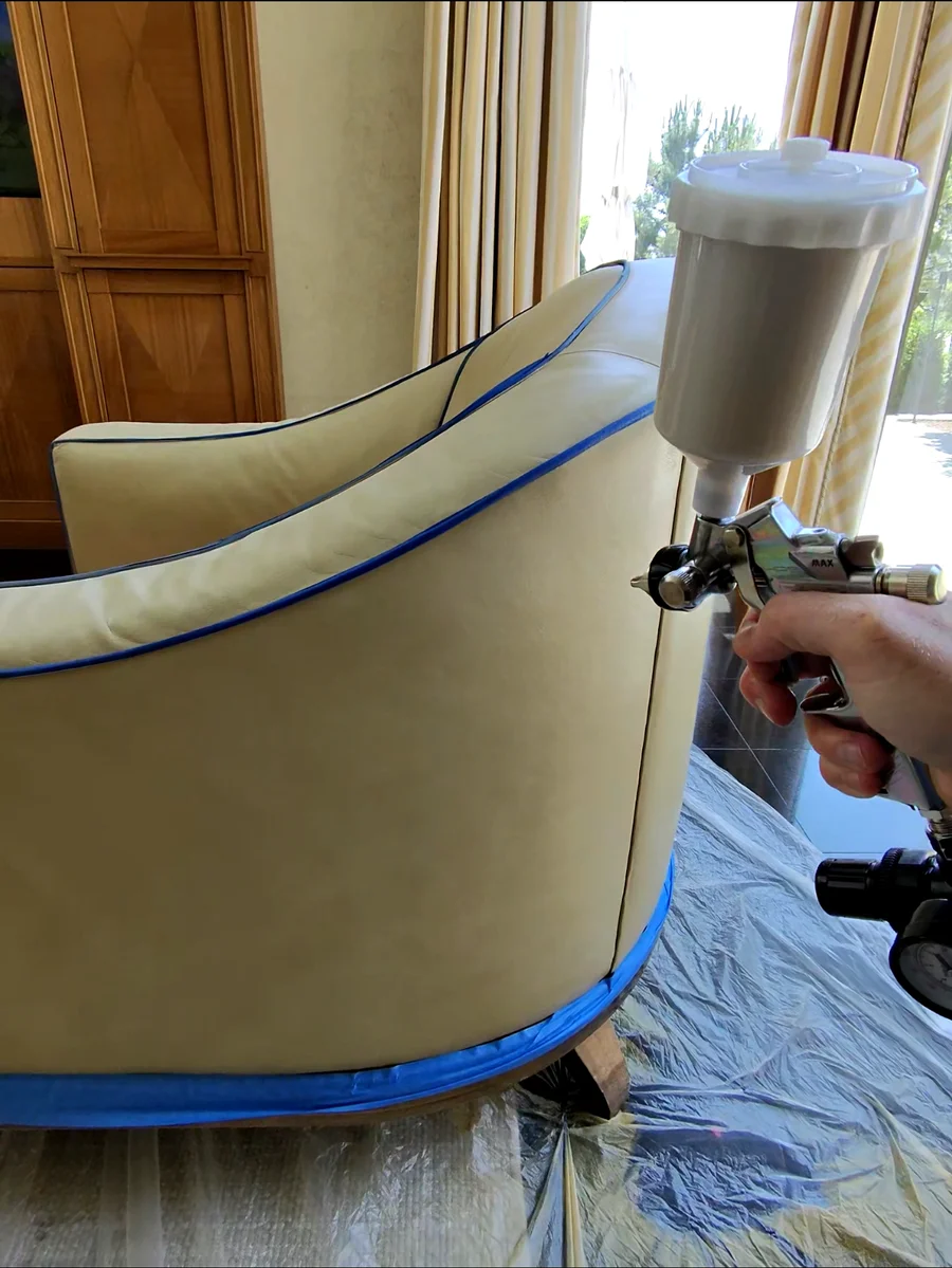 Professional leather color application on club chair — blue masking tape protects white contrast piping while spray color is applied during luxury furniture restoration