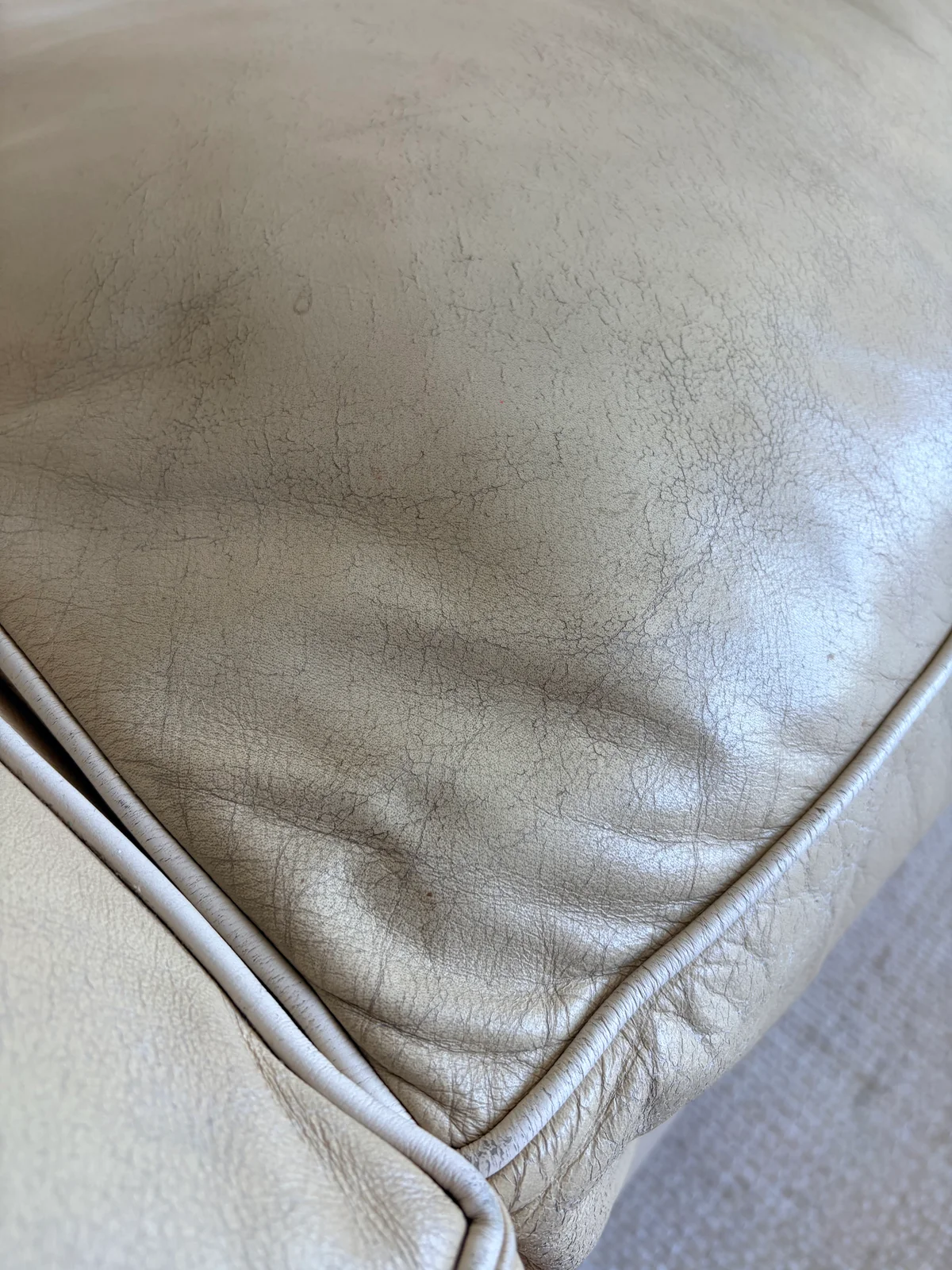 Close-up of severe leather finish failure and cracking on sofa cushion corner — extensive crack network visible across UV-damaged surface with white contrast piping